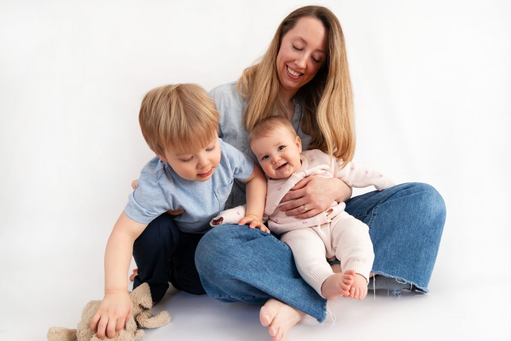 Older brother sitting with baby sister in a studio, smiling and cuddling, neutral white background family portrait