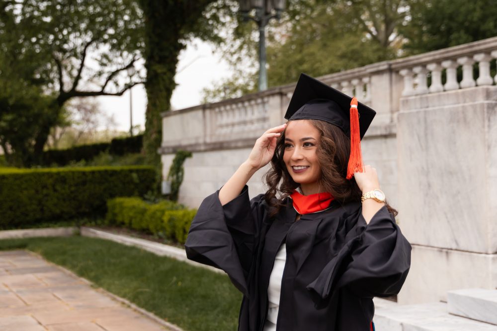 Smiling graduate in black cap and gown with red hood during a graduation photoshoot in Cleveland, Ohio, posing outdoors on a grassy campus