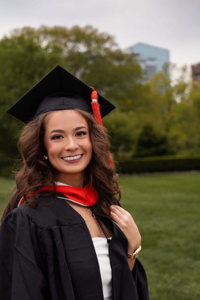 Smiling graduate in black cap and gown with red hood during a graduation photoshoot in Cleveland, Ohio, posing outdoors on a grassy campus