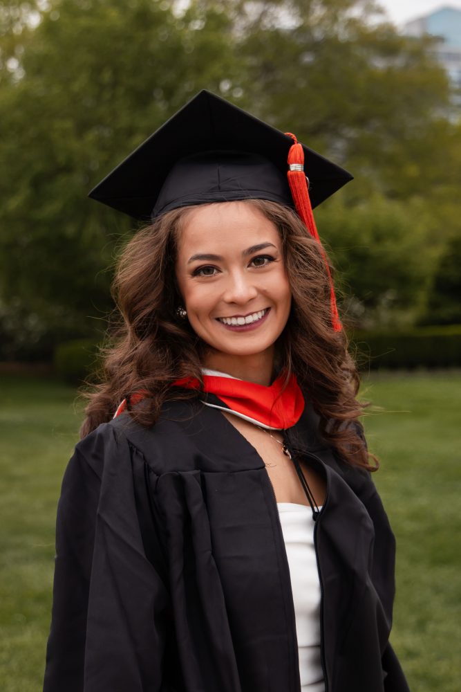 Smiling graduate in black cap and gown with red hood during a graduation photoshoot in Cleveland, Ohio, posing outdoors on a grassy campus