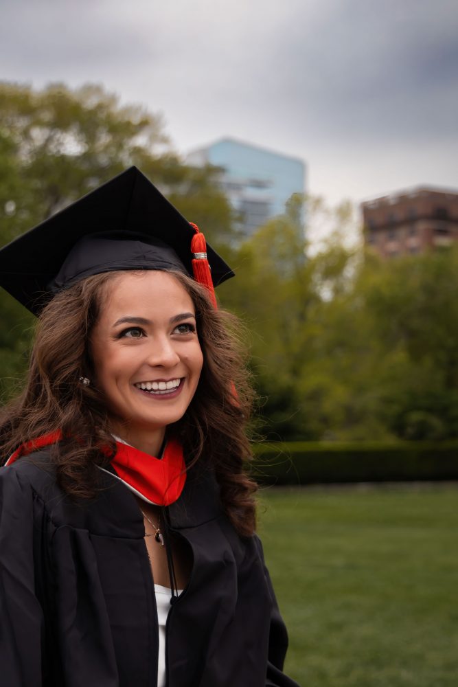 Smiling graduate in black cap and gown with red hood during a graduation photoshoot in Cleveland, Ohio, posing outdoors on a grassy campus