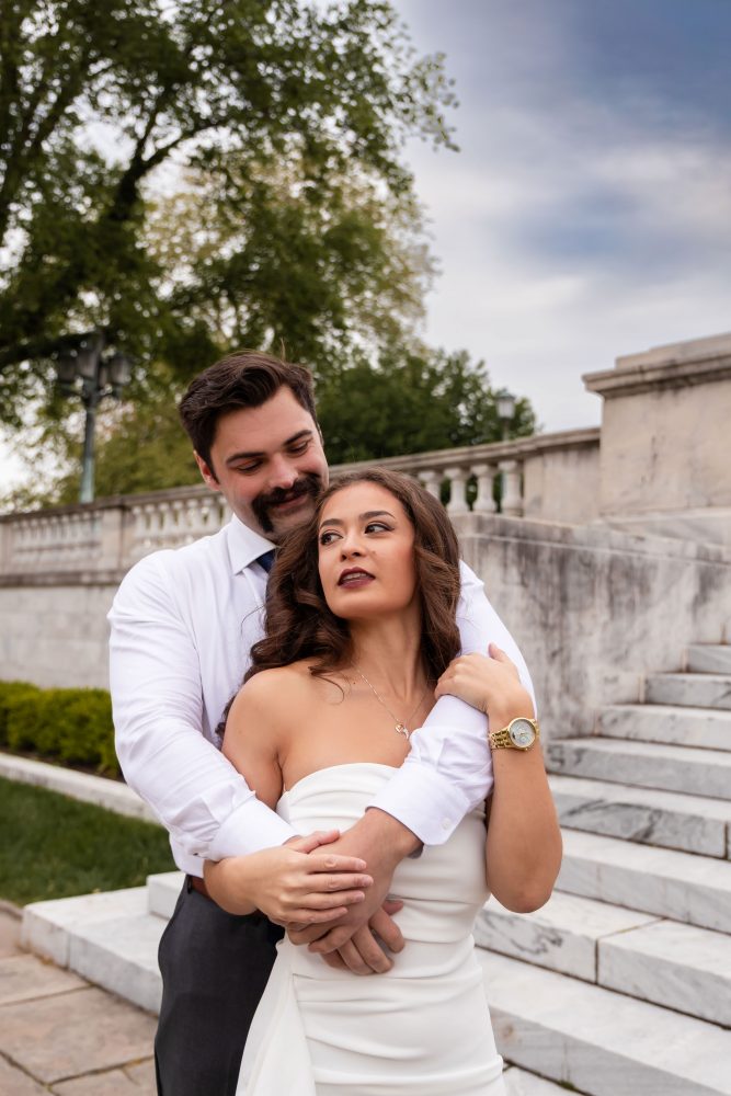 Couple embracing near marble steps during a graduation photoshoot in Cleveland, Ohio, with the woman in a white dress looking over her shoulder while the man smiles behind her