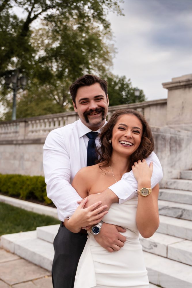 Couple embracing near marble steps during a graduation photoshoot in Cleveland, Ohio, with the woman in a white dress looking over her shoulder while the man smiles behind her