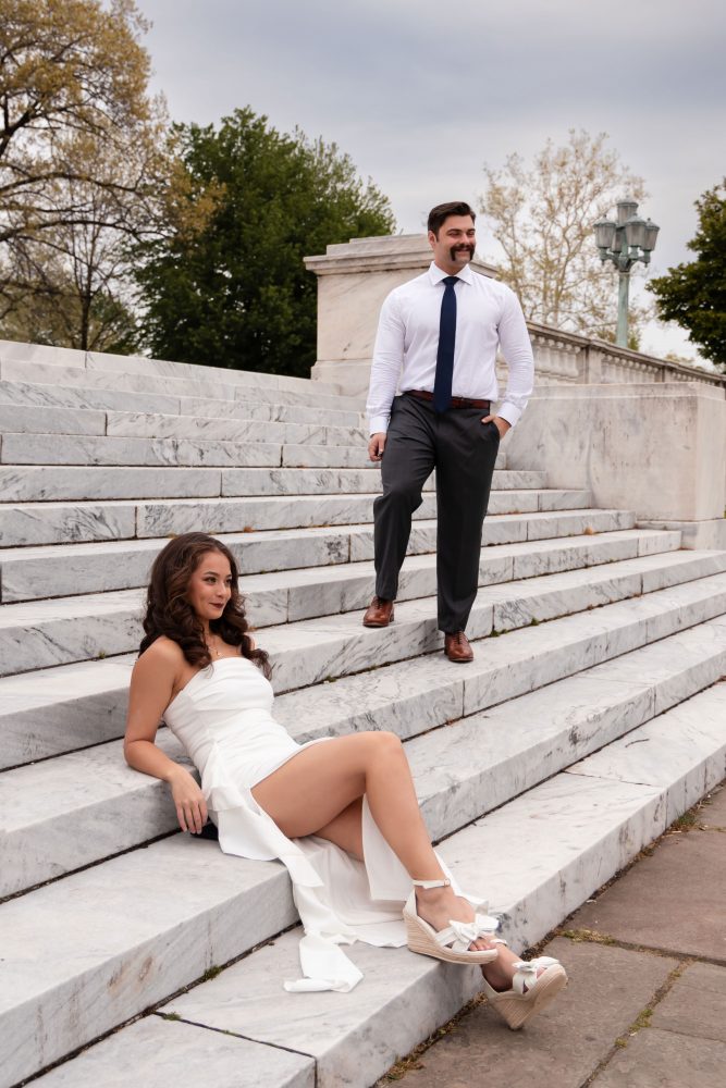 Couple embracing near marble steps during a graduation photoshoot in Cleveland, Ohio, with the woman in a white dress looking over her shoulder while the man smiles behind her