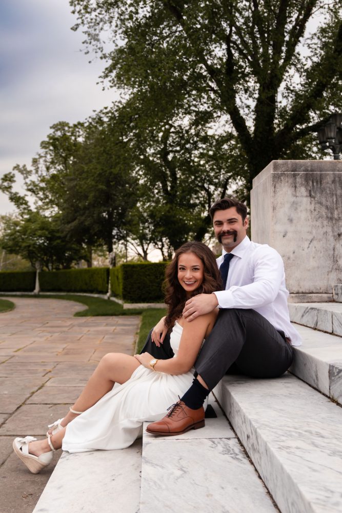 Couple embracing near marble steps during a graduation photoshoot in Cleveland, Ohio, with the woman in a white dress looking over her shoulder while the man smiles behind her