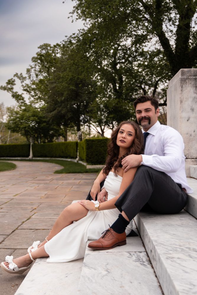 Couple embracing near marble steps during a graduation photoshoot in Cleveland, Ohio, with the woman in a white dress looking over her shoulder while the man smiles behind her
