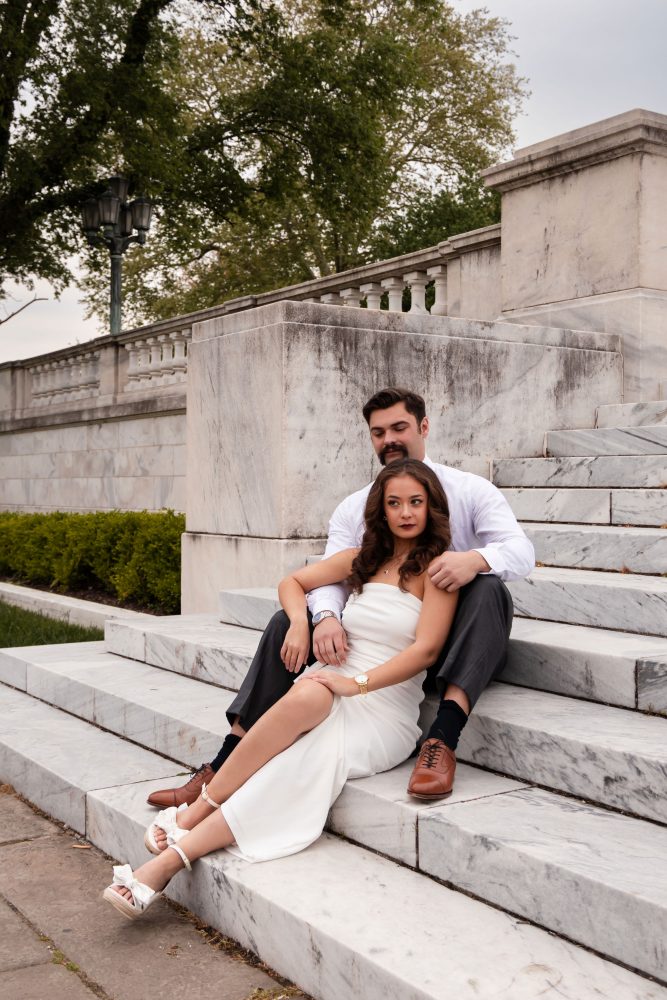 Couple embracing near marble steps during a graduation photoshoot in Cleveland, Ohio, with the woman in a white dress looking over her shoulder while the man smiles behind her