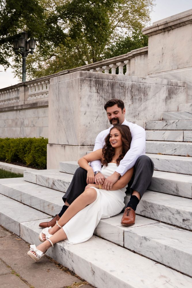 Couple embracing near marble steps during a graduation photoshoot in Cleveland, Ohio, with the woman in a white dress looking over her shoulder while the man smiles behind her