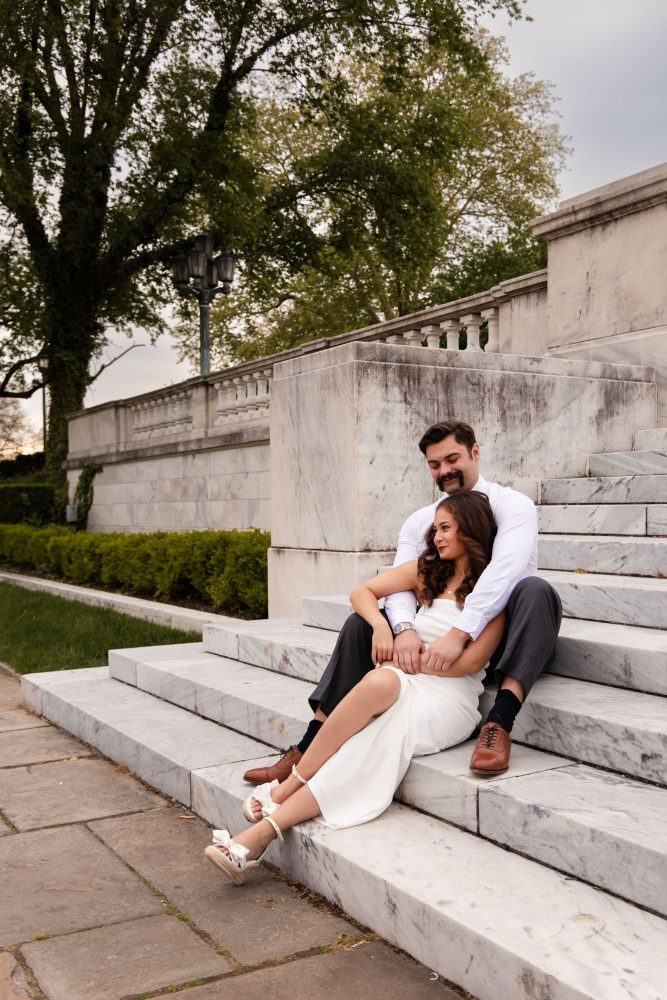 Couple embracing near marble steps during a graduation photoshoot in Cleveland, Ohio, with the woman in a white dress looking over her shoulder while the man smiles behind her