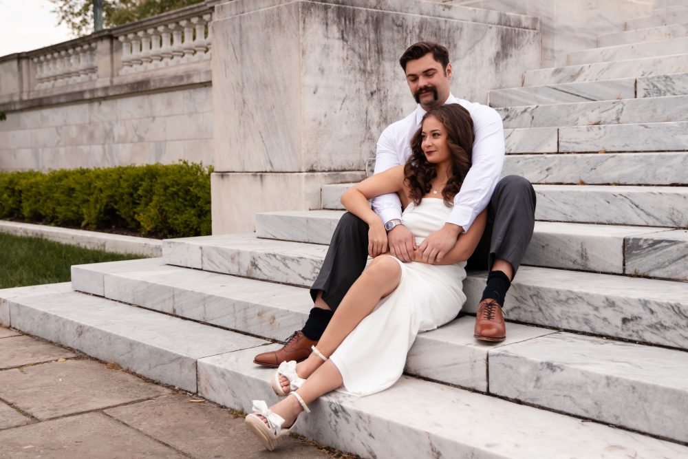 Couple embracing near marble steps during a graduation photoshoot in Cleveland, Ohio, with the woman in a white dress looking over her shoulder while the man smiles behind her