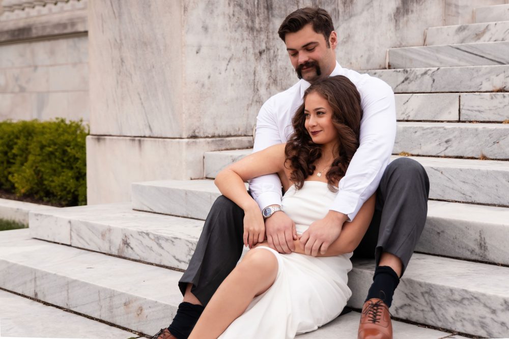 Couple embracing near marble steps during a graduation photoshoot in Cleveland, Ohio, with the woman in a white dress looking over her shoulder while the man smiles behind her