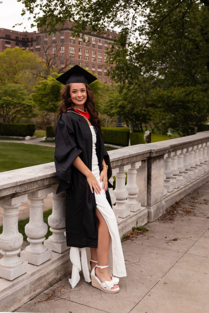 Smiling graduate in black cap and gown with red hood during a graduation photoshoot in Cleveland, Ohio, posing outdoors on a grassy campus.