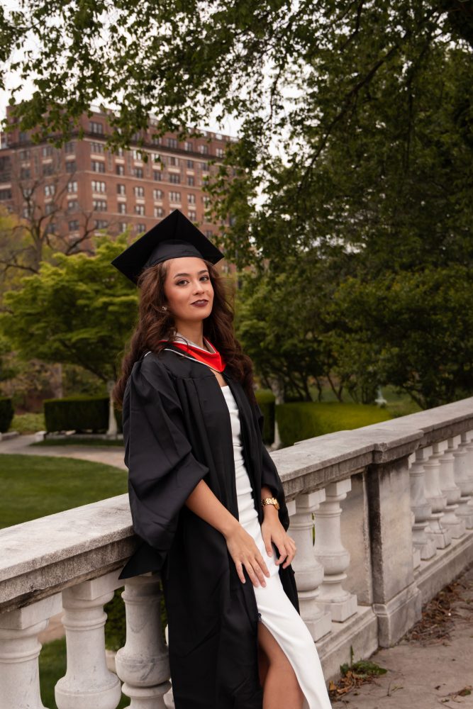 Smiling graduate in black cap and gown with red hood during a graduation photoshoot in Cleveland, Ohio, posing outdoors on a grassy campus.