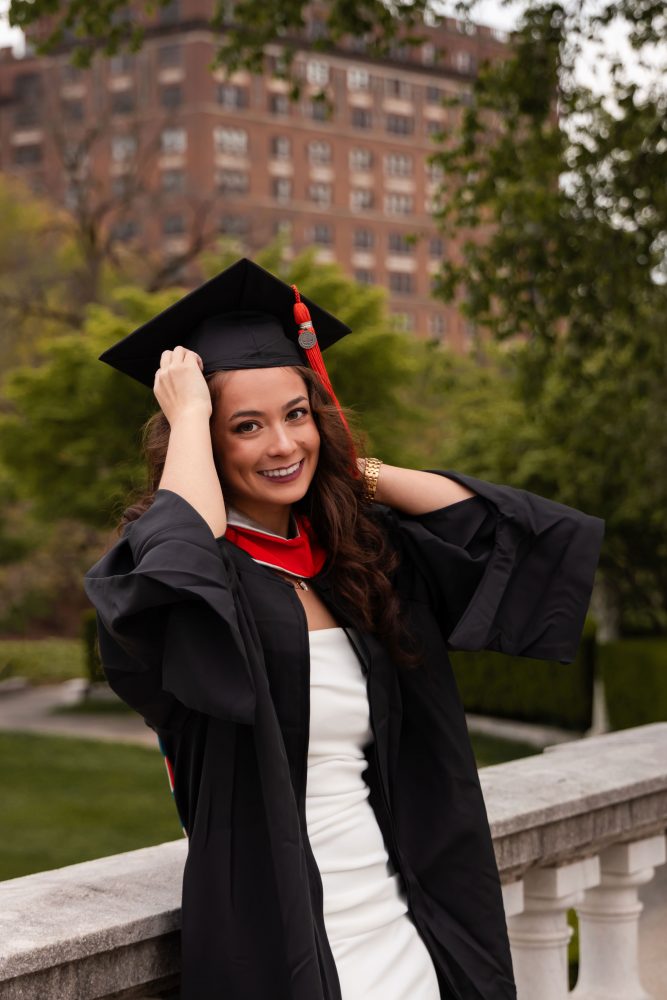 Smiling graduate in black cap and gown with red hood during a graduation photoshoot in Cleveland, Ohio, posing outdoors on a grassy campus.