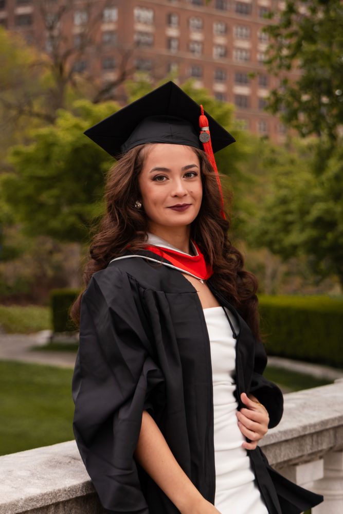 Smiling graduate in black cap and gown with red hood during a graduation photoshoot in Cleveland, Ohio, posing outdoors on a grassy campus.