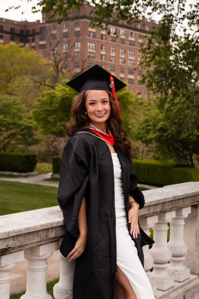 Smiling graduate in black cap and gown with red hood during a graduation photoshoot in Cleveland, Ohio, posing outdoors on a grassy campus.