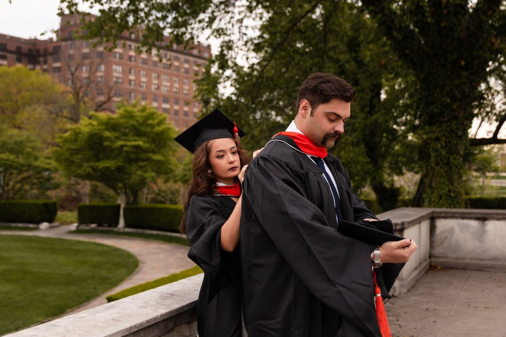 Smiling graduate in black cap and gown with red hood during a graduation photoshoot in Cleveland, Ohio, posing outdoors on a grassy campus.