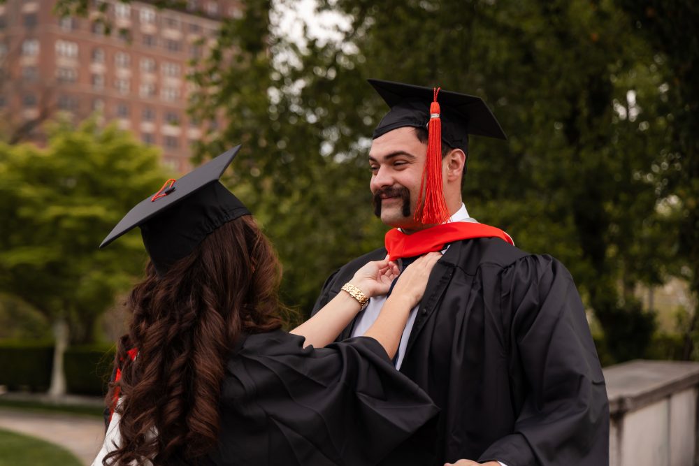 Smiling graduate in black cap and gown with red hood during a graduation photoshoot in Cleveland, Ohio, posing outdoors on a grassy campus.
