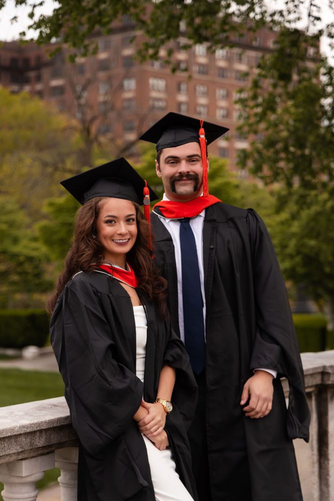 Smiling graduate in black cap and gown with red hood during a graduation photoshoot in Cleveland, Ohio, posing outdoors on a grassy campus.