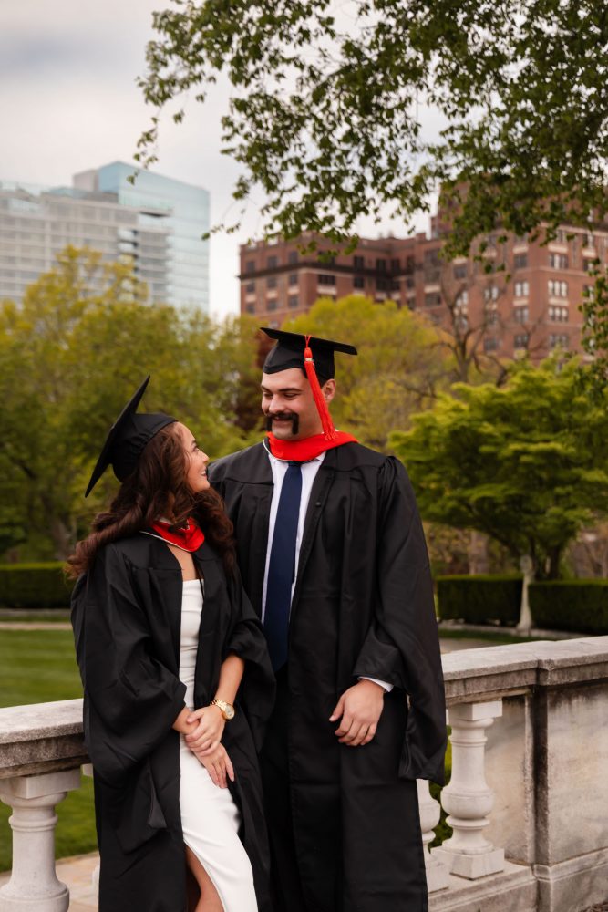 Smiling graduate in black cap and gown with red hood during a graduation photoshoot in Cleveland, Ohio, posing outdoors on a grassy campus.