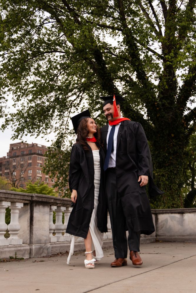Smiling graduate in black cap and gown with red hood during a graduation photoshoot in Cleveland, Ohio, posing outdoors on a grassy campus.