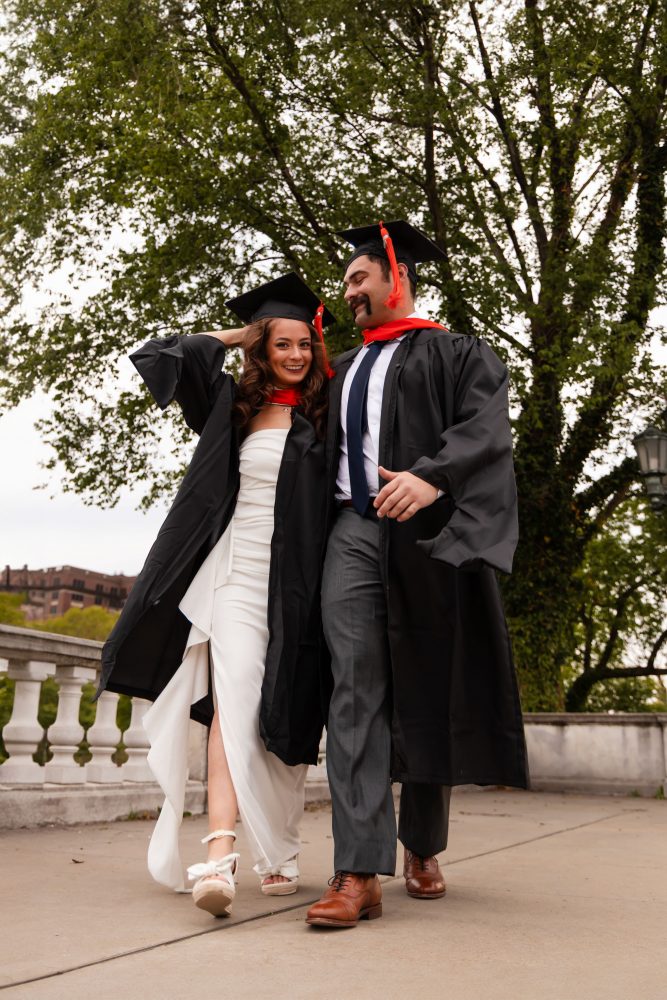 Smiling graduate in black cap and gown with red hood during a graduation photoshoot in Cleveland, Ohio, posing outdoors on a grassy campus.