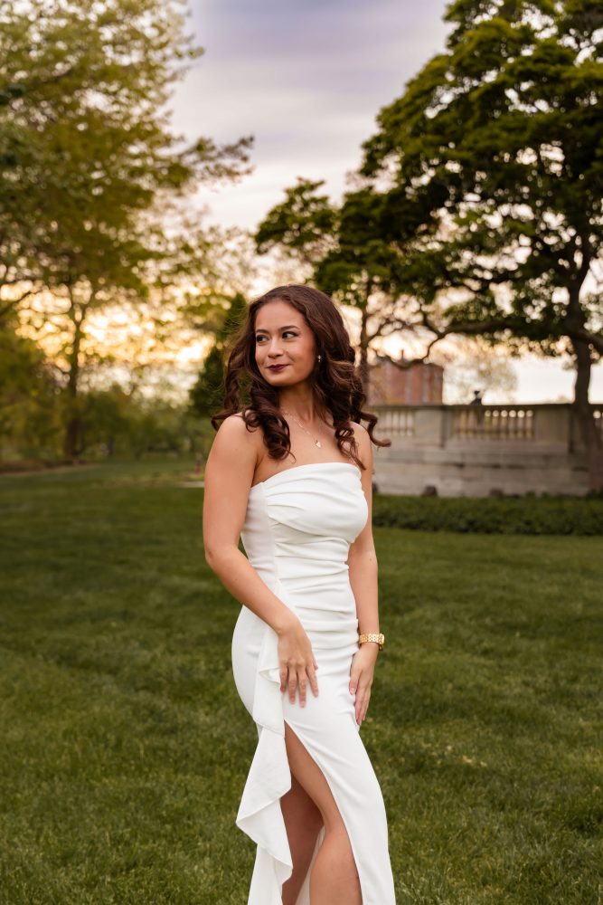 Young woman in an elegant white dress posing in a park at sunset during a graduation photoshoot in Cleveland, Ohio, with soft golden light and trees in the background