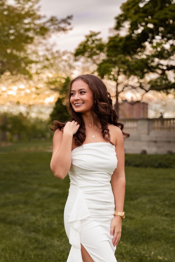 Young woman in an elegant white dress posing in a park at sunset during a graduation photoshoot in Cleveland, Ohio, with soft golden light and trees in the background