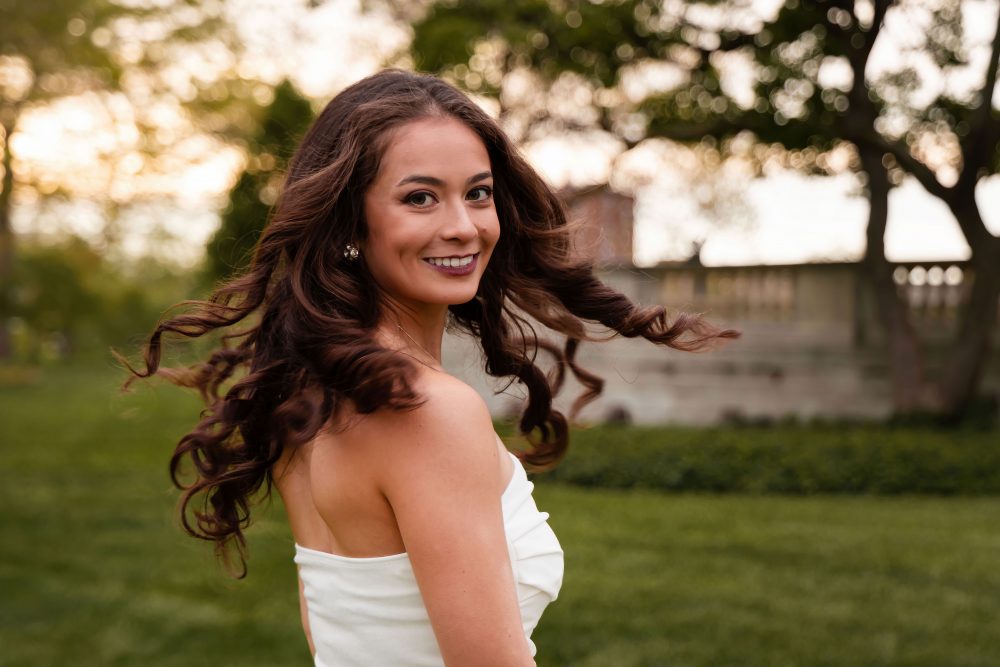 Young woman in an elegant white dress posing in a park at sunset during a graduation photoshoot in Cleveland, Ohio, with soft golden light and trees in the background