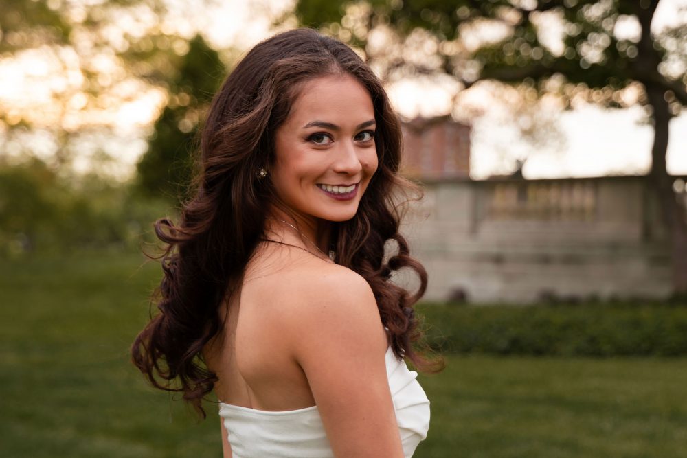 Young woman in an elegant white dress posing in a park at sunset during a graduation photoshoot in Cleveland, Ohio, with soft golden light and trees in the background
