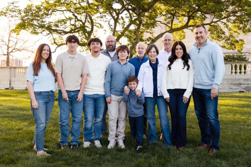 Ten members of an extended family, including grandparents, adults, teenagers, and a young child, walk together across a grassy lawn outside the Cleveland Museum of Art. The group smiles and looks at one another as the museum building and trees appear in the background.
