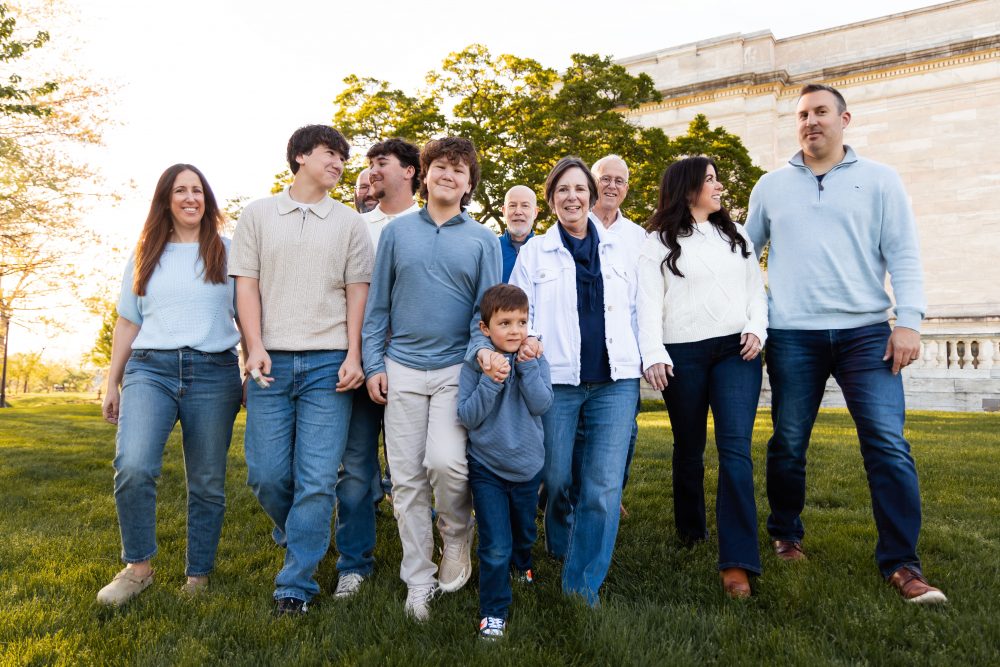 Ten members of an extended family, including grandparents, adults, teenagers, and a young child, walk together across a grassy lawn outside the Cleveland Museum of Art. The group smiles and looks at one another as the museum building and trees appear in the background.