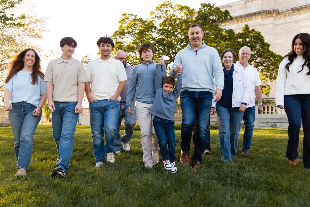 Ten members of an extended family, including grandparents, adults, teenagers, and a young child, walk together across a grassy lawn outside the Cleveland Museum of Art. The group smiles and looks at one another as the museum building and trees appear in the background.