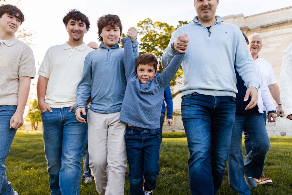 Ten members of an extended family, including grandparents, adults, teenagers, and a young child, walk together across a grassy lawn outside the Cleveland Museum of Art. The group smiles and looks at one another as the museum building and trees appear in the background.