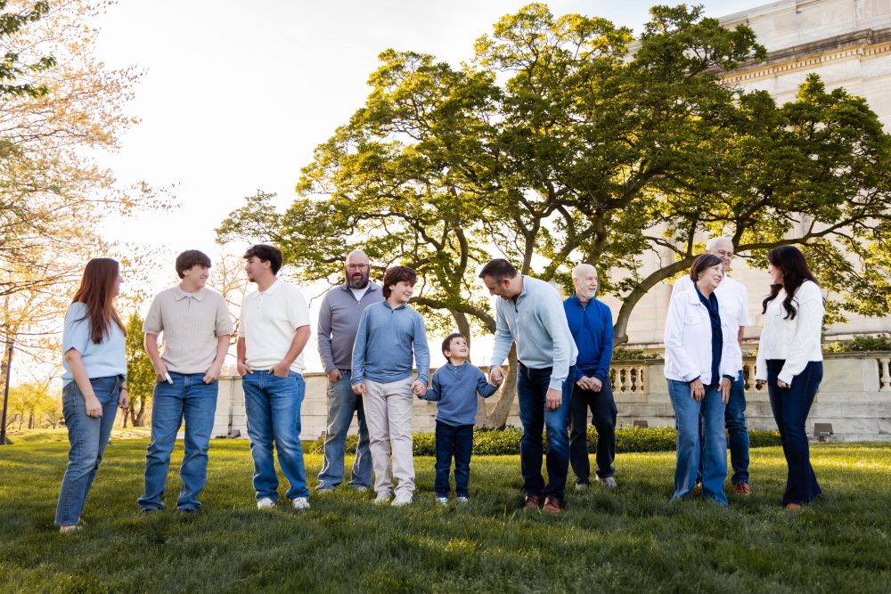 Ten members of an extended family, including grandparents, adults, teenagers, and a young child, walk together across a grassy lawn outside the Cleveland Museum of Art. The group smiles and looks at one another as the museum building and trees appear in the background.