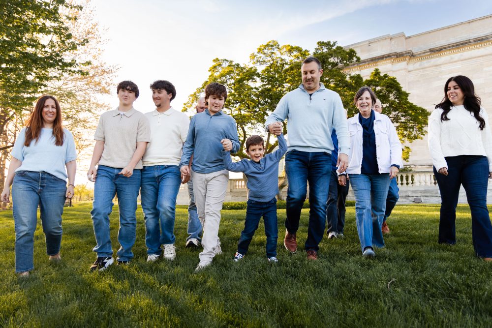 Ten members of an extended family, including grandparents, adults, teenagers, and a young child, walk together across a grassy lawn outside the Cleveland Museum of Art. The group smiles and looks at one another as the museum building and trees appear in the background.