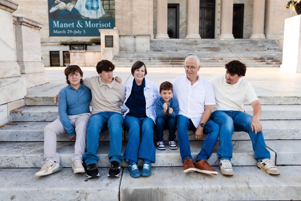 Ten members of an extended family, including grandparents, adults, teenagers, and a young child, walk together across a grassy lawn outside the Cleveland Museum of Art. The group smiles and looks at one another as the museum building and trees appear in the background.
