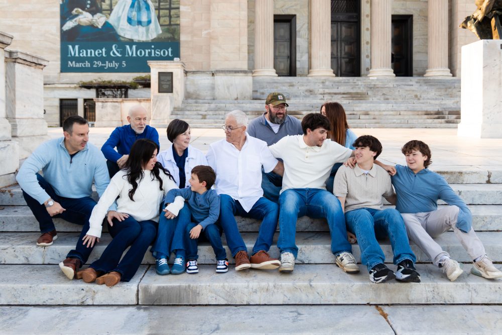 Ten members of an extended family, including grandparents, adults, teenagers, and a young child, walk together across a grassy lawn outside the Cleveland Museum of Art. The group smiles and looks at one another as the museum building and trees appear in the background.