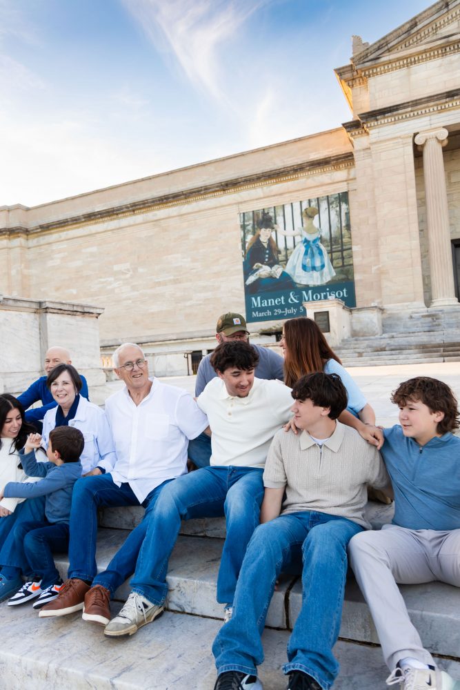 Ten members of an extended family, including grandparents, adults, teenagers, and a young child, walk together across a grassy lawn outside the Cleveland Museum of Art. The group smiles and looks at one another as the museum building and trees appear in the background.