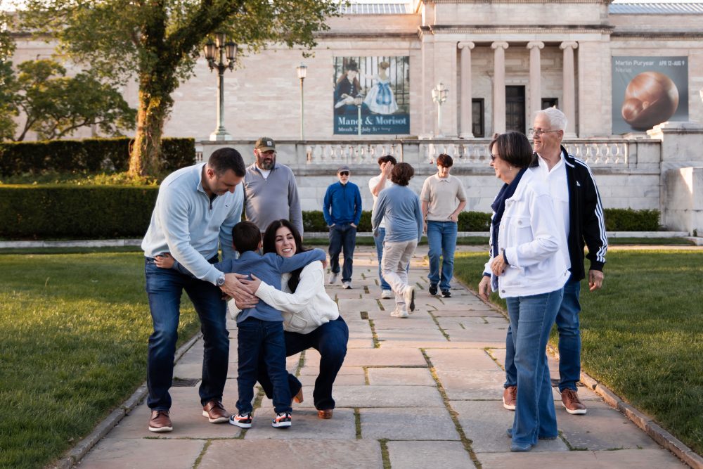 Ten members of an extended family, including grandparents, adults, teenagers, and a young child, walk together across a grassy lawn outside the Cleveland Museum of Art. The group smiles and looks at one another as the museum building and trees appear in the background.