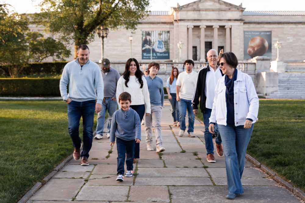 Ten members of an extended family, including grandparents, adults, teenagers, and a young child, walk together across a grassy lawn outside the Cleveland Museum of Art. The group smiles and looks at one another as the museum building and trees appear in the background.