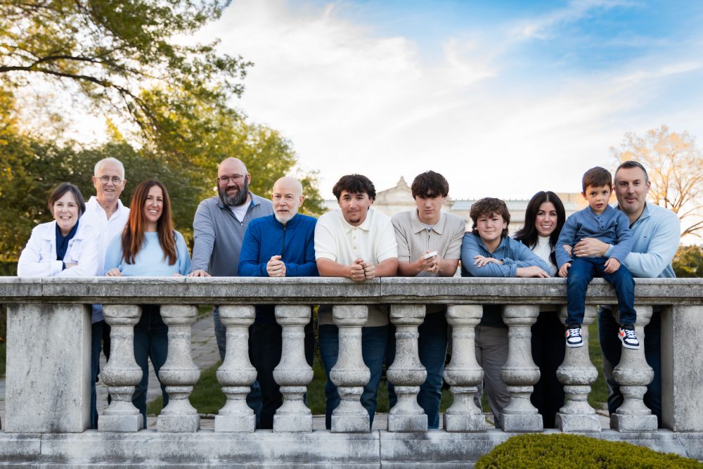 Ten members of an extended family, including grandparents, adults, teenagers, and a young child, walk together across a grassy lawn outside the Cleveland Museum of Art. The group smiles and looks at one another as the museum building and trees appear in the background.