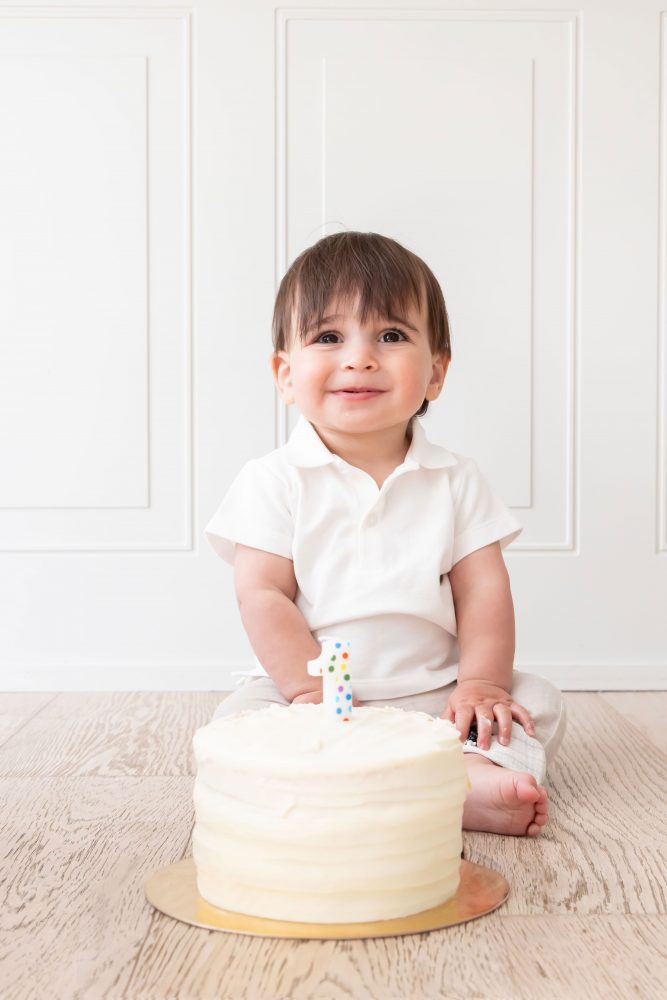 One-year-old baby boy sitting beside a vanilla birthday cake during a classic first birthday cake smash session in Cleveland, Ohio.