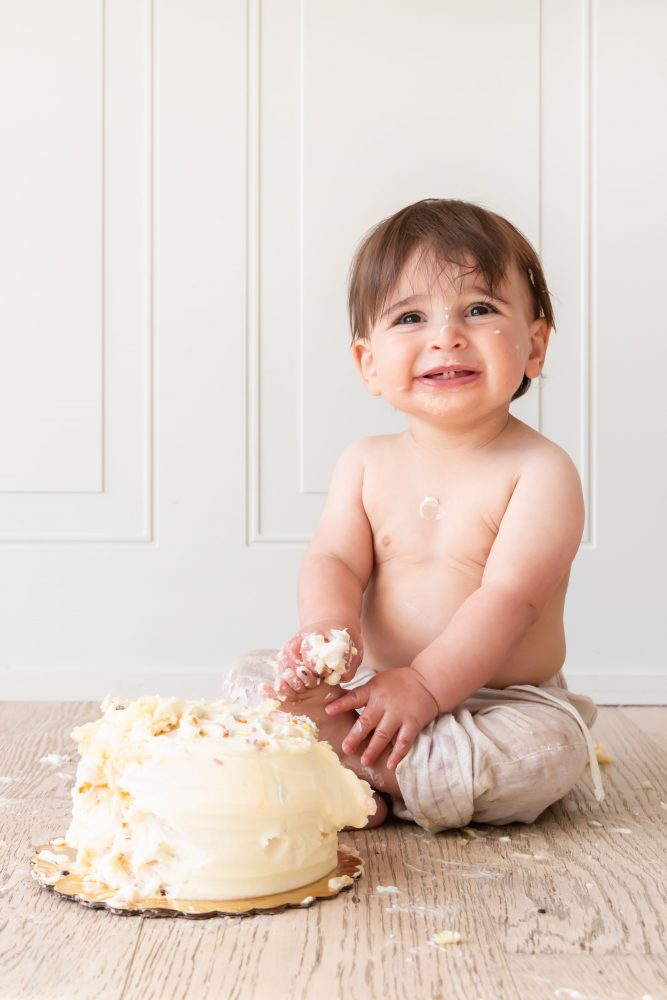 First birthday cake smash photography of a smiling baby boy with frosting-covered hands in Cleveland, Ohio.