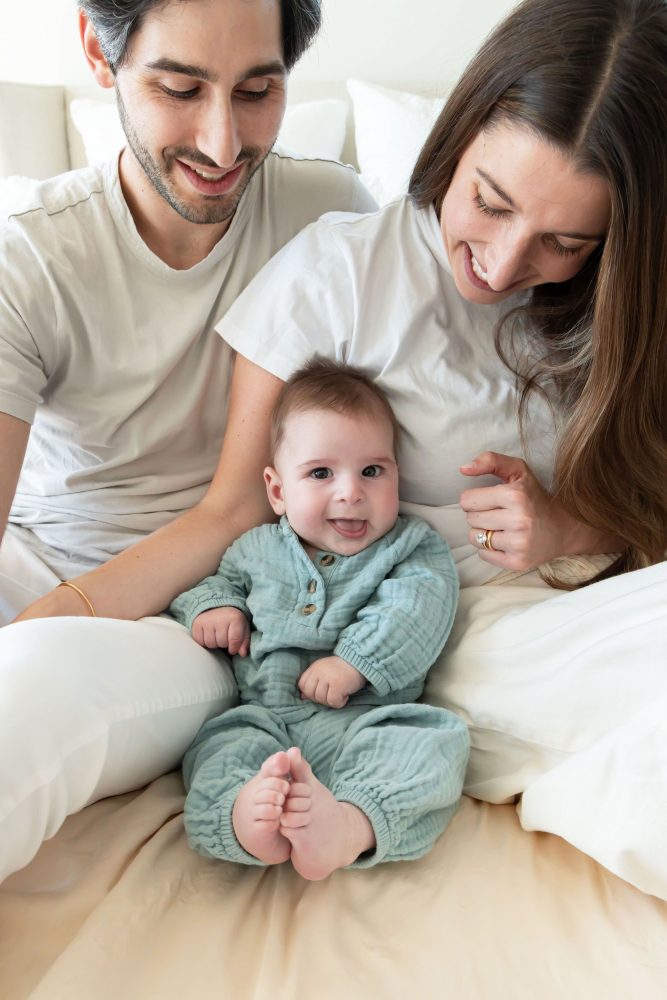 Lifestyle newborn family session in a bright bedroom by a Cleveland family photographer, with parents cuddling their smiling baby on a white bed.
