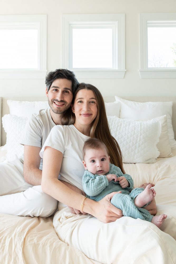 Lifestyle newborn family session in a bright bedroom by a Cleveland family photographer, with parents cuddling their smiling baby on a white bed.