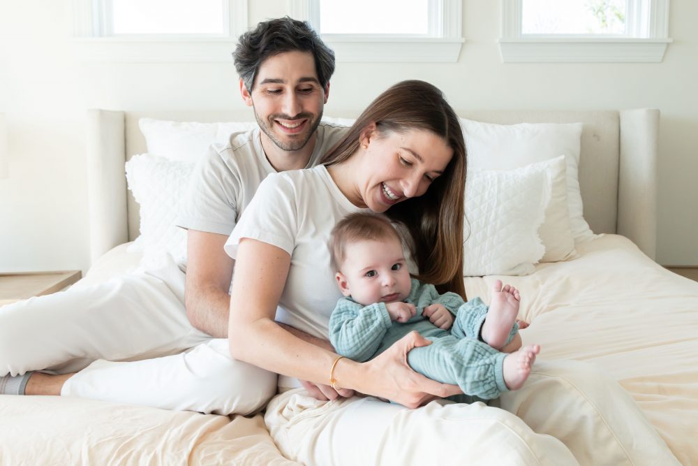 Lifestyle newborn family session in a bright bedroom by a Cleveland family photographer, with parents cuddling their smiling baby on a white bed.