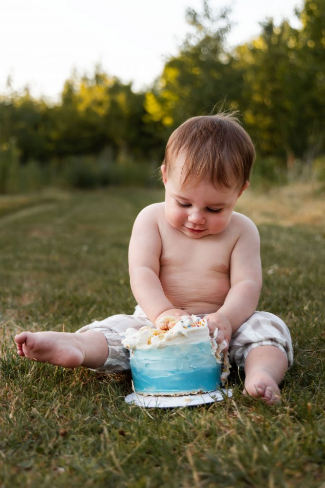 Baby boy celebrating his first birthday with frosting on his cheeks and hands during a playful outdoor cake smash.
