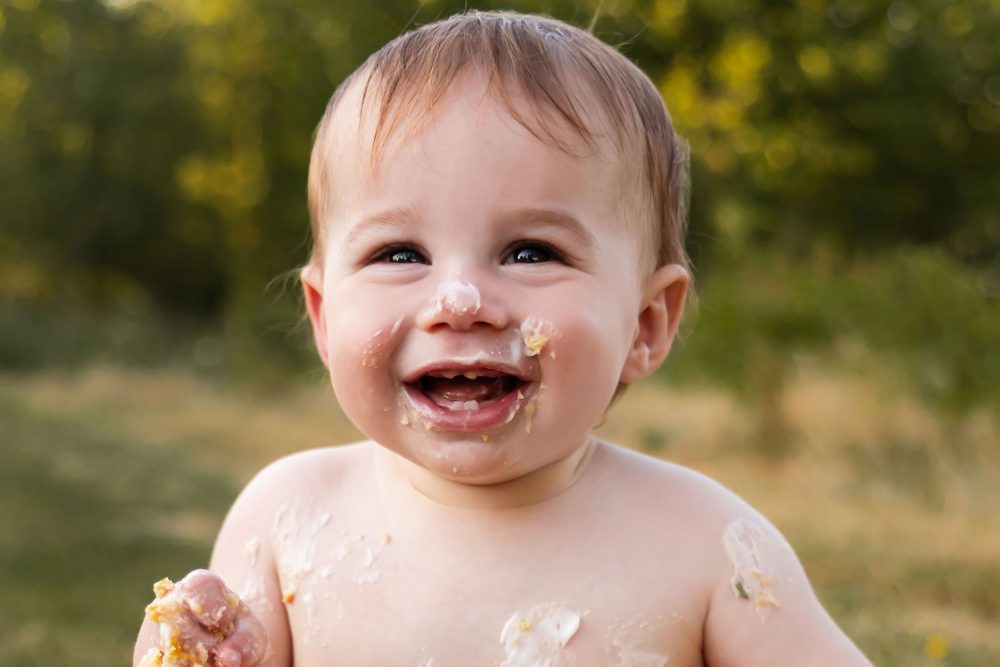Outdoor first birthday cake smash portrait featuring a smiling baby boy sitting beside his smashed birthday cake in Cleveland, Ohio.