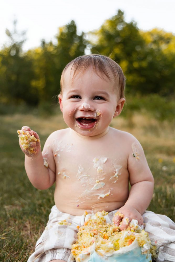 Outdoor first birthday cake smash portrait featuring a smiling baby boy sitting beside his smashed birthday cake in Cleveland, Ohio.